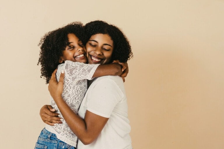 A young girl wearing a white lace top and jeans hugs a woman in a white t-shirt. Both are smiling with eyes closed, standing against a plain beige background, capturing the essence of heartwarming daughter quotes.