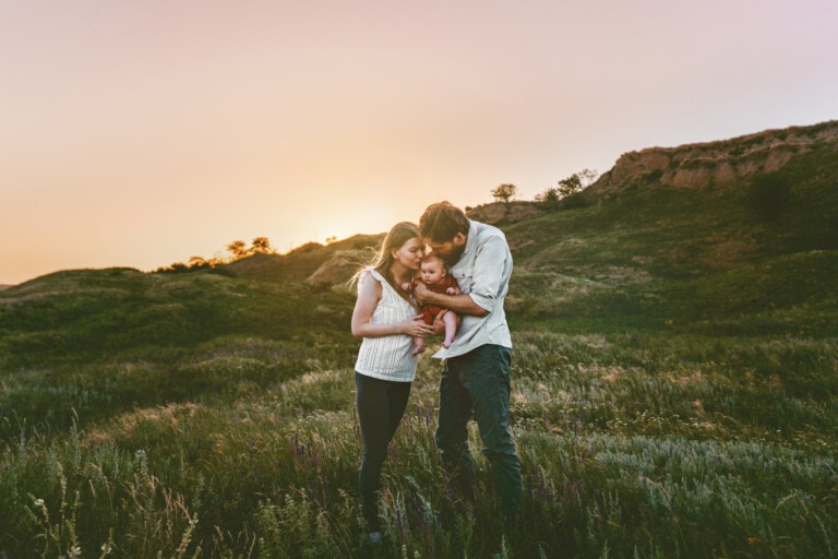 A couple stands in a grassy field at sunset, holding a baby. The mother wears a white sleeveless top and black pants, while the father wears a light-colored shirt and dark pants. The father kisses the baby on the head as they all pose together, savoring less alone time but more cherished moments.