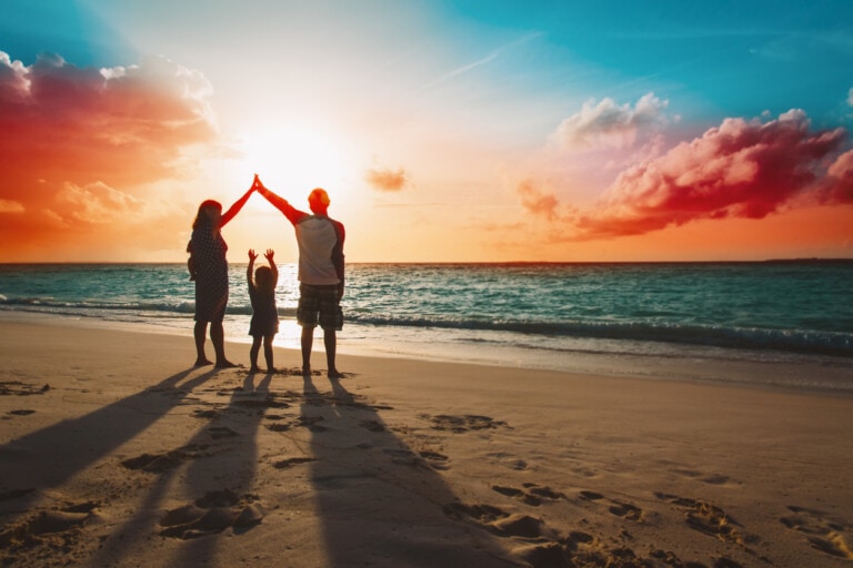 A family of four stands on a beach at sunset, enjoying one of their memorable vacations. The parents and two children hold hands, forming arches with their arms. The sun sets over the ocean in the background, casting long shadows on the sand. Pink clouds and a mix of blue and orange hues fill the sky.