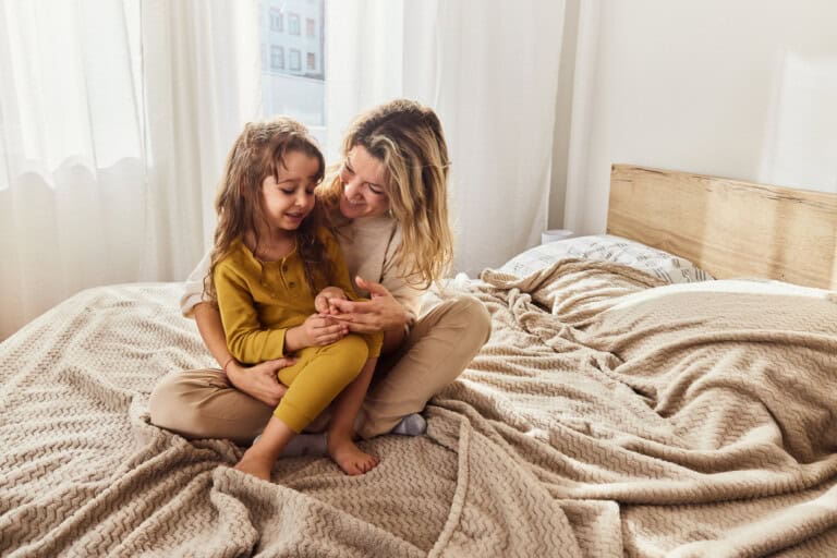 A woman and a child sit on an unmade bed with beige blankets and a wooden headboard. The woman has her arms around the child and they are both smiling. Sunlight streams through a window with sheer curtains on the left.