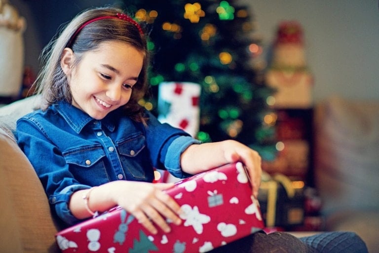 A young girl with a red headband and blue shirt is sitting on a couch, smiling as she holds one of the best gifts for little girls: a red gift-wrapped present. In the background, there is a decorated Christmas tree with lights and ornaments.