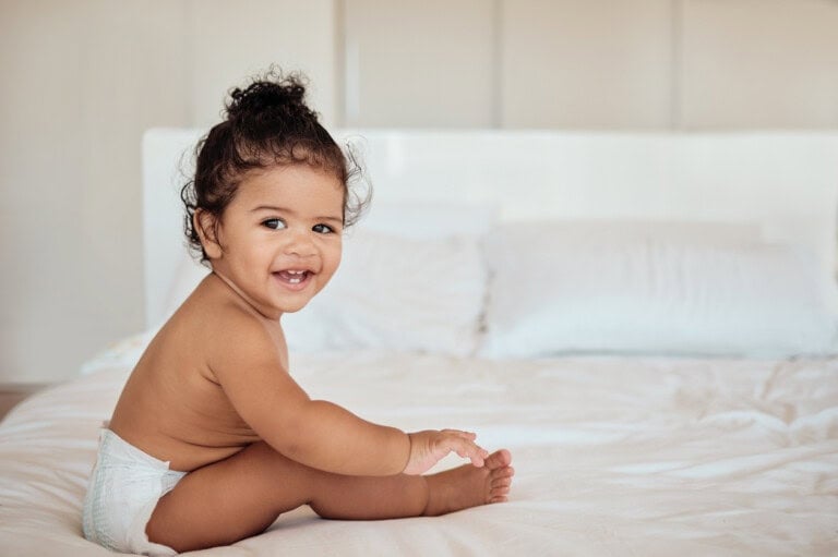 A 15-month-old baby wearing a diaper is sitting on a white bed. The baby is smiling and looking towards the camera. The room has a bright and neutral appearance with white pillows and bedding in the background.