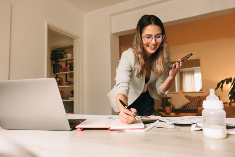 A woman wearing glasses is standing at a desk, holding a phone and writing in a notebook. There is a laptop, papers, and a baby bottle on the desk. A living room is visible in the background, suggesting she’s setting boundaries to manage her work-life balance.