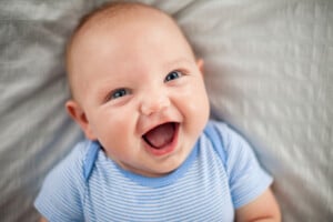A baby boy with blue eyes and light hair is lying on a white blanket, wearing a light blue and white striped onesie, and smiling with an open mouth. Parents looking for boy names that start with J might find inspiration from his joyful expression.