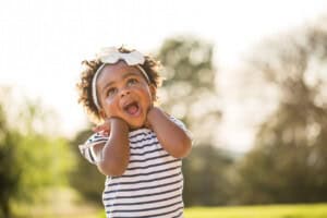 A 14-month-old toddler with curly hair and a white bow is standing outside with greenery in the background. The child, dressed in a striped shirt, has an open-mouthed expression while holding their hands near their face, possibly curious about new activities for 14-month-olds.
