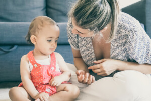 A baby sits on the floor wearing a red dress with white polka dots. An adult with a patterned top and tied-back hair is sitting next to the baby, looking and gesturing towards them, perhaps trying to get kids to listen. They are in a room with a grey couch in the background.