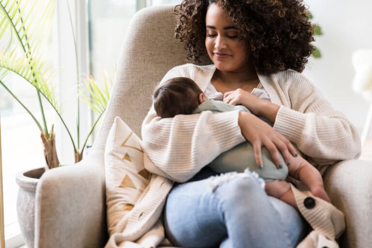 A woman with curly hair is sitting in a comfortable chair near a window, cradling her baby. She gazes down lovingly, wearing a light-colored cardigan and jeans. A lush plant adds a touch of greenery in the background.