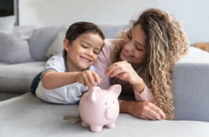 A young boy and a woman with curly hair lie on a couch, smiling as they place coins into a pink piggy bank. Several coins are scattered next to it, illustrating an early lesson in budgeting. The background features a white wall and a gray couch.