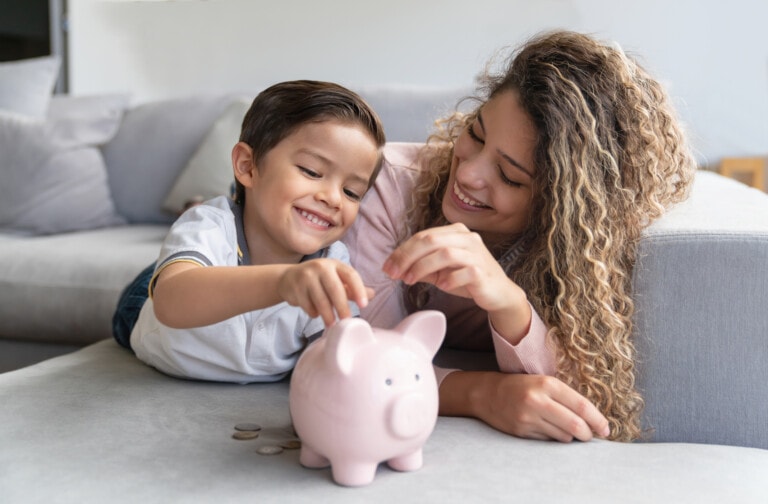 A young boy and a woman with curly hair lie on a couch, smiling as they place coins into a pink piggy bank. Several coins are scattered next to it, illustrating an early lesson in budgeting. The background features a white wall and a gray couch.