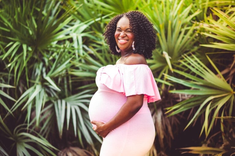 A pregnant woman smiles while posing outdoors in front of lush greenery. She is wearing a pink off-the-shoulder dress, one of the chic maternity summer clothes options, and has one hand resting on her belly.