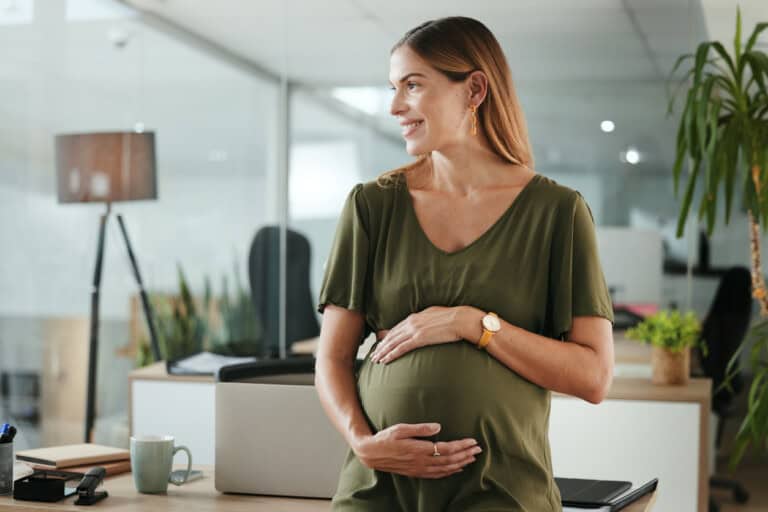 A pregnant woman in a green dress stands in an office setting, smiling and cradling her pregnant belly. She is leaning against a desk with office supplies. There are plants and modern furniture in the background.