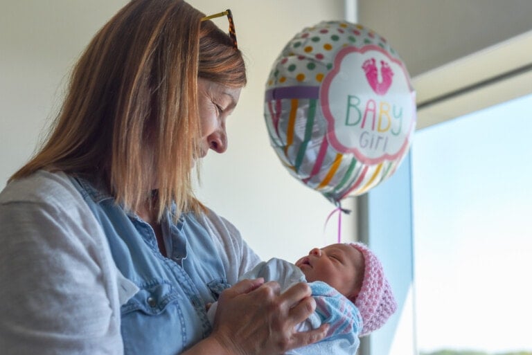 A woman holds a sleeping newborn wrapped in a blanket while looking at the baby lovingly. A "Baby Girl" balloon floats in the background, hinting that people visit your newborn to share in the joyous moment. The room is well-lit with natural light coming through a window.