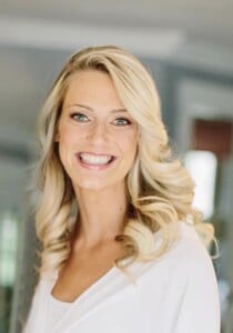 A woman with long, wavy blonde hair is smiling broadly. She is wearing a white top, and the background is slightly blurred, suggesting an indoor setting.