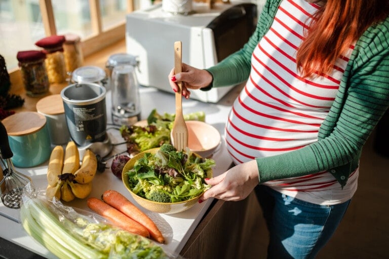A pregnant woman with red hair in a striped shirt and green cardigan is mixing a salad in a bowl in her kitchen, focusing on foods to eat in the third trimester. The counter is filled with various vegetables like bananas, carrots, and celery. In the background, jars and kitchen appliances are visible.
