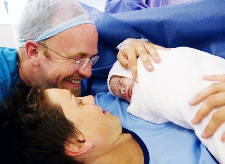 A newborn baby wrapped in a white blanket is being cradled by someone lying on a hospital bed after an unexpected c-section, while another person, wearing a surgical cap and smiling, looks on closely. The scene appears to be in a delivery room.