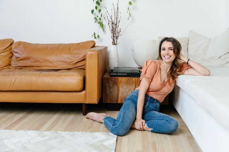 A woman in a casual outfit of a peach top and blue jeans is sitting on the floor, leaning against a white couch. Behind her, there is a tan leather sofa, a wooden side table with books, and a vase with branches and green leaves. The space is minimally decorated, creating an atmosphere close to respectful baby sleep conditions.