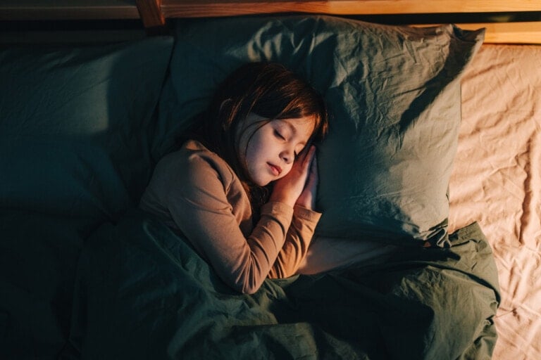 A young child with long dark hair is sleeping peacefully in a bed with green sheets and a pillow. The child is lying on their side with hands tucked under the face. The room is dimly lit, filled with the gentle hum of white noise, creating a calm and serene atmosphere.