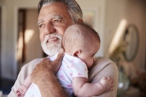 An older man with gray hair and a beard, perhaps affectionately known by one of many grandpa names, holds a baby wearing a white outfit with pink designs. The man is looking off to the side while the baby rests on his shoulder. The background is indoors, featuring a mirror and various household items.