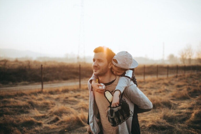 A man is walking outdoors with a child on his back, showcasing one of the many ways dads show their love. The child, wearing a baseball cap backward, holds a baseball glove. They pass by a wire fence with a field and utility poles in the background as the sunlight casts a warm glow.