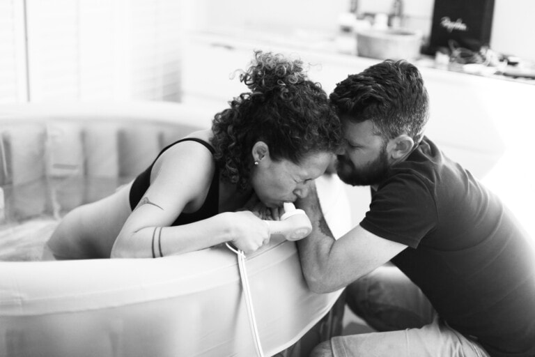 A black and white image of a woman in a birthing pool, gripping the edge and leaning forward. A man sits beside her, his forehead against hers in support. In the home setting, you can see the subtle presence of nitrous oxide equipment nearby, hinting at its use for pain relief.