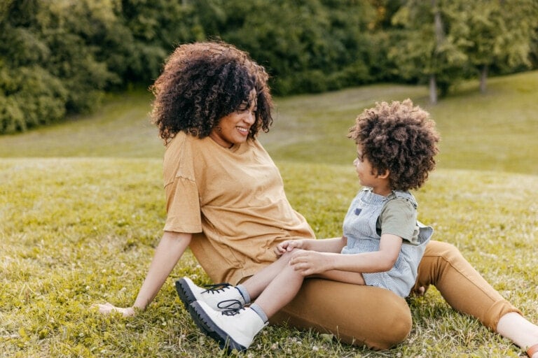 A person with curly hair is sitting on the grass, leaning back on one arm, and smiling at a child with curly hair who is sitting on their lap. As they enjoy the serene background of grassy fields and trees, they share loving phrases to say to your child.