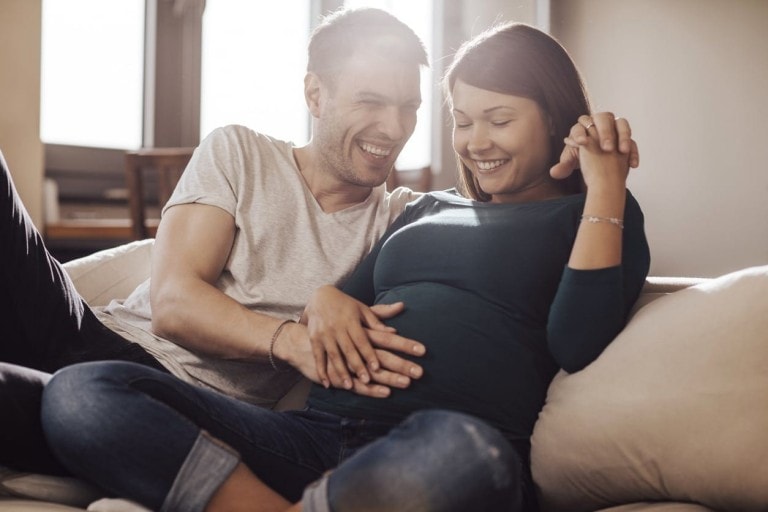 A couple sits on a couch, smiling and looking at each other. The woman, wearing a long-sleeved green shirt, holds her pregnant belly with one hand, while her other hand is held by the man in a short-sleeved gray shirt. They appear happy and relaxed as they discuss things to do before baby arrives.