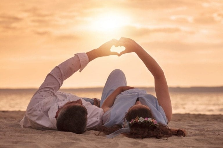 A couple lying on the sand at the beach during sunset, taking a babymoon. They are facing each other and forming a heart shape with their hands towards the sun, which is low in the sky. The woman, who is on vacation while pregnant, wears a blue dress and a floral headband, while the man is in a white shirt.