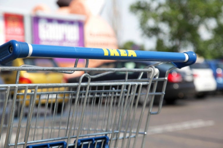 A close-up of an IKEA shopping cart handle in a parking lot. The blue handle bears the yellow IKEA logo. In the background, several finds at IKEA blur into view among parked cars and some visible green foliage.