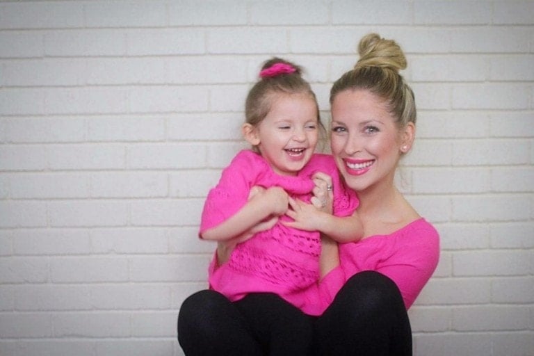 A woman and a young girl, both with blonde hair in a bun and wearing bright pink tops, are sitting against a white brick wall. The woman is holding the laughing girl on her lap, both smiling as if sharing secrets about their favorite essential oils for naptime.