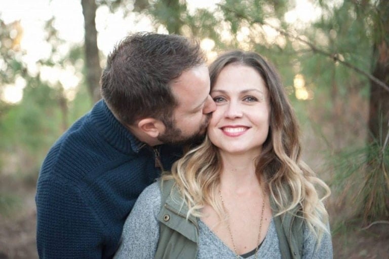 A man with short hair and a beard kisses a smiling woman with long, wavy hair on the cheek. They are outdoors, surrounded by trees, with blurred greenery in the background. The man wears a dark sweater, and the woman wears a gray top with a green vest—a testament to their strong marriage.