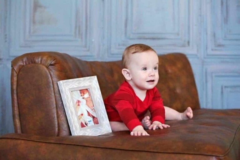 A baby wearing a red outfit sits on a brown leather couch with a framed photo placed beside them. The background consists of a light blue wall with decorative molding. The neonatal ICU graduate is looking towards the right, appearing curious.