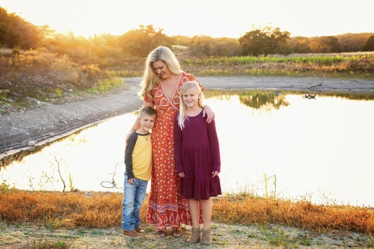 A widow in a red patterned dress stands next to a boy in a yellow shirt and a girl in a purple dress by a pond. The sun is setting behind them, casting a warm glow over the scene. They are standing on grass with water and trees in the background.