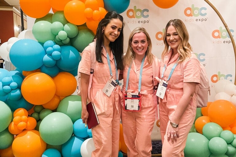 Three women wearing matching pink jumpsuits stand in front of a colorful balloon arrangement and an "abc kids expo" banner. Each holds event badges and smiles at the camera, showcasing the best of ABC kids expo.