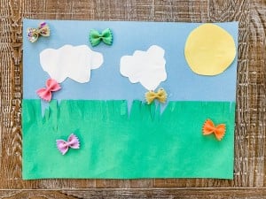 A children's artwork shows a green grassy field under a blue sky with two white clouds and a yellow sun. Colorful bowtie pasta pieces, painted in various colors, are glued onto the paper as flowers or butterflies, making this one of the easy crafts for kids. The background is a wooden surface.