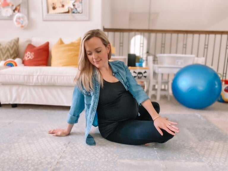 A pregnant woman with long blonde hair is sitting on the floor in a living room, possibly preparing for some pregnancy stretches. She is wearing a black top and dark pants with a blue denim shirt. Behind her are a white couch with colorful pillows, a large blue exercise ball, and a baby activity seat.