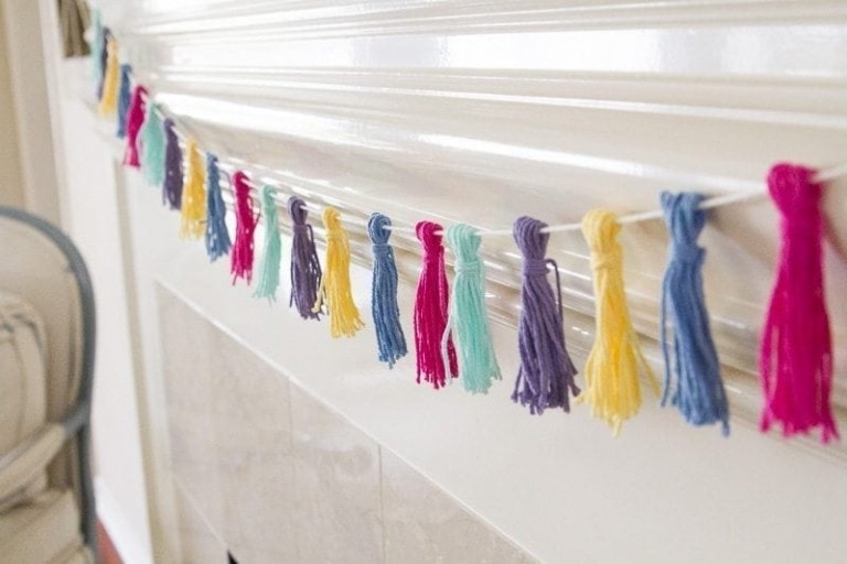 A close-up of a spring tassel garland hanging as a festive touch on what appears to be a white mantelpiece. The multicolored tassels in blue, purple, pink, yellow, green, and turquoise bring vibrant charm to the décor.