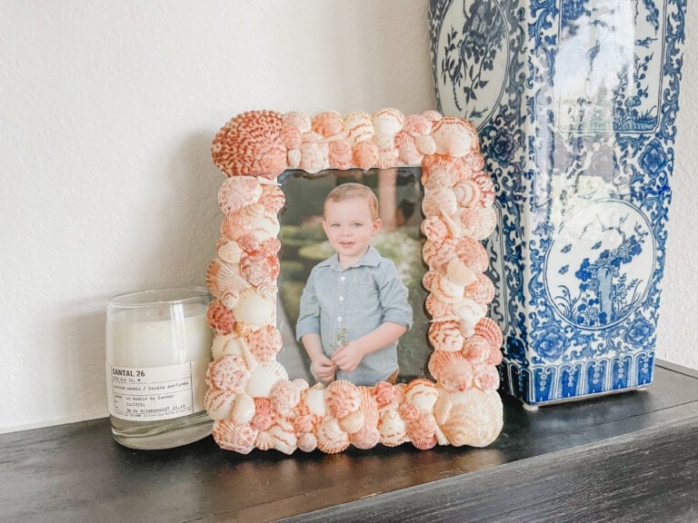 A DIY shell frame holds a picture of a young child in a blue shirt, standing. The frame is placed on a black table next to a white candle in a glass jar labeled "SANTAL 26" and a blue and white decorative vase.