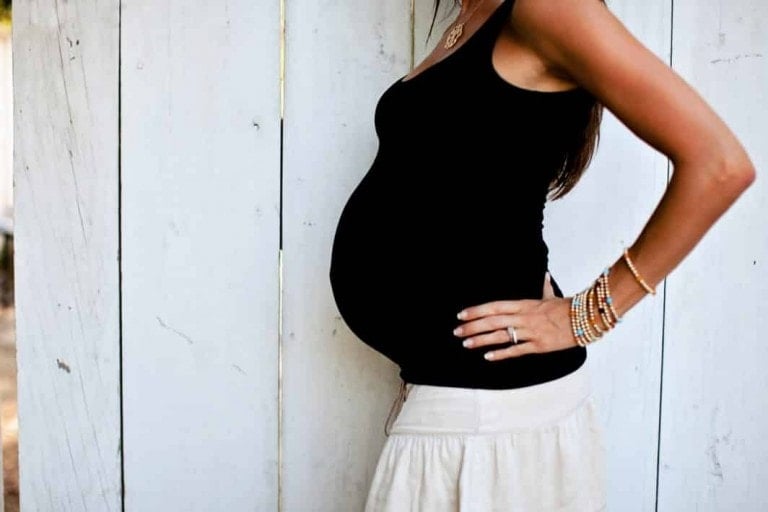 A pregnant person stands sideways with hands on hips, pondering what pregnancy cravings mean. They are wearing a black tank top, a cream-colored skirt, and multiple bracelets on their wrist. The background is a white wooden wall.