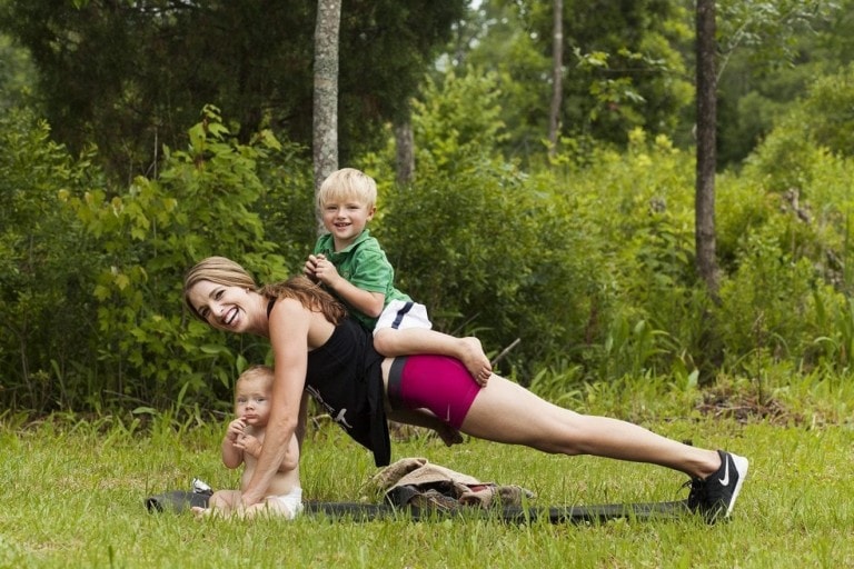 A woman performs a plank exercise on a yoga mat outdoors, smiling warmly. In this joyful postpartum workout, a child in a green shirt lies on her back and holds onto her neck, while another child sits on the grass beside her. Trees and greenery fill the background.