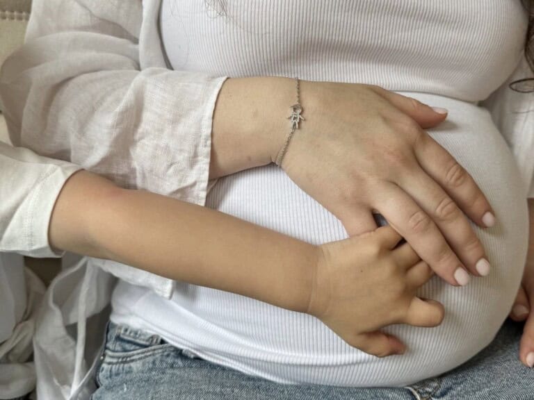 A close-up image shows an adults hand and a childs hand gently resting on a pregnant womans belly. The adult wears a white long-sleeve shirt, a silver Oliviar bracelet, and blue jeans. The child, in a white shirt, embraces the belly, indicating a nurturing gesture.