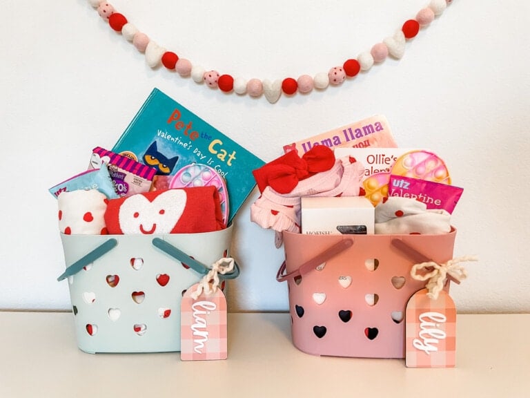 Two Valentine's Day gift baskets, one light blue and one pink, filled with children's books, toys, and cloth items. Each basket has a tag with a name and heart-shaped cutouts. A string of pink and white puffballs hangs on the wall behind them.
