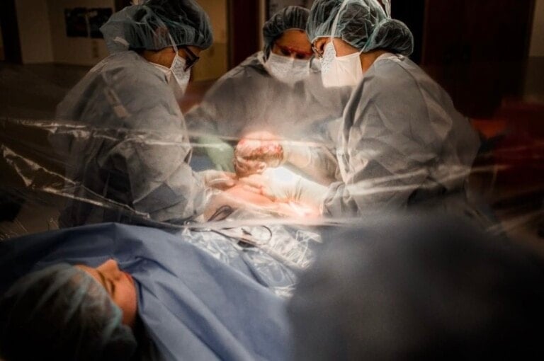 Medical professionals in surgical attire, including masks and scrub hats, perform a gentle cesarean on a patient lying on an operating table. One doctor holds a newborn baby, with a protective plastic sheet partially covering the patient.