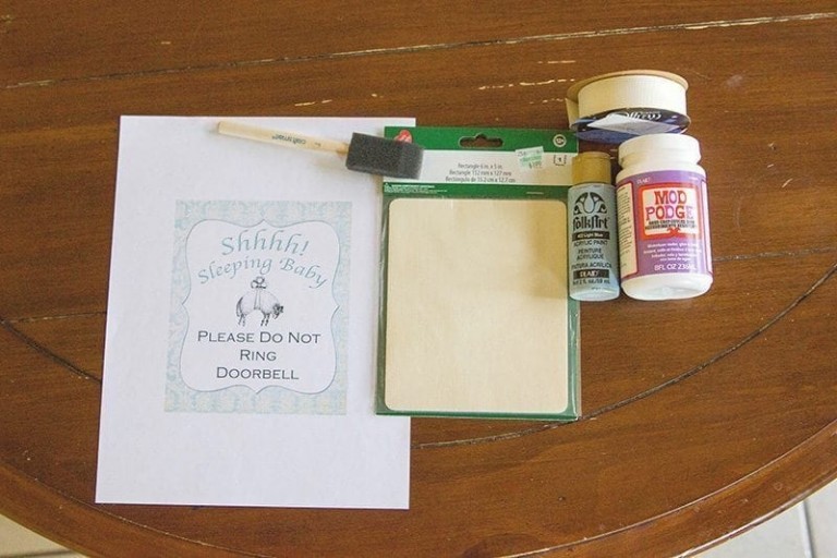 A wooden table displays a printed "Do Not Disturb Baby" sign, a sponge brush, a small wooden board, a bottle of white paint, a jar of glitter, and a container of Mod Podge.