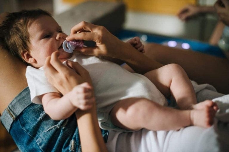 An adult is holding a baby who is lying on their lap, gently placing a pacifier into the baby's mouth. The baby, in a white onesie, seems content. The background appears to be a home setting with a sofa and some out-of-focus elements, symbolizing the daily pros of pacifiers in soothing infants.