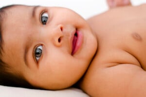 A close-up image of a baby lying down, looking slightly to the side. The baby, possibly on the brink of being given one of those beautiful Indian names for girls, has large, dark eyes and a small open mouth. The baby appears to be partially undressed, showing their bare upper body. The background is out of focus and light-colored.