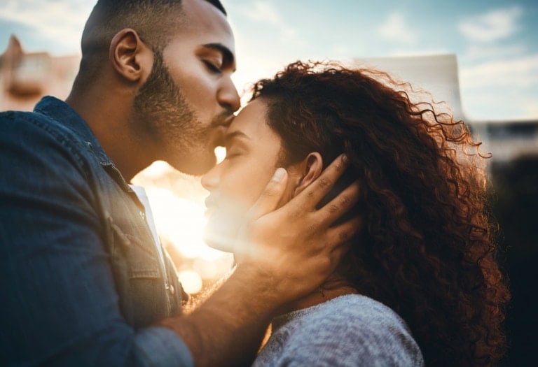 A man gently kisses a woman on the forehead as he cups her face with his hand. Their eyes are closed, appearing serene and wrapped in a moment of pure intimacy. The background is slightly blurred, highlighting them against a sunset, casting an ethereal glow that makes their love feel alive in parenthood.