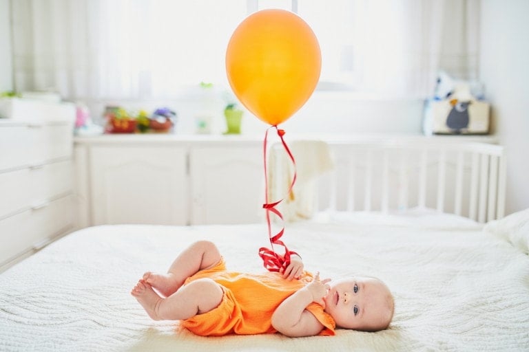 A baby dressed in an orange outfit lies on a bed holding the string of an orange balloon, as if ready for their Sip and See. The room is well-lit with natural light coming from the window in the background, highlighting the white furniture and decor.