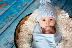A baby is wrapped in a light blue blanket and wearing a light blue knit hat. The baby is lying in a round wooden basket lined with fluffy white fabric against a background of weathered blue wooden planks, perhaps dreaming of c names for boys like Charles or Caleb, with one hand near the mouth.