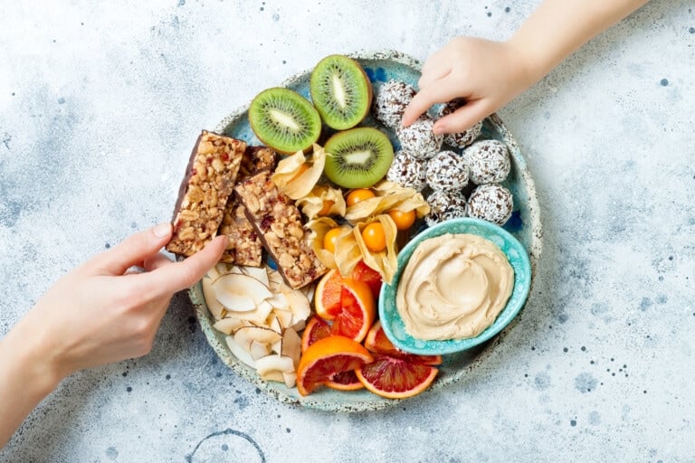 A plate with an assortment of healthy snacks for kids, including granola bars, sliced kiwi, dried fruit, a small bowl of dip, and energy balls covered in coconut flakes. Two hands are reaching for food from the plate. The background is a light speckled surface.