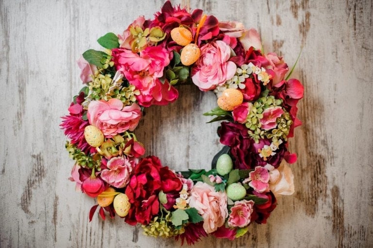 A spring wreath with vibrant pink, red, and green artificial flowers and foliage, adorned with small decorative Easter eggs in pastel colors, is placed against a light wooden background.